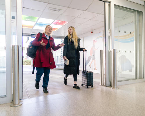 Two women with luggage walking through some double doors